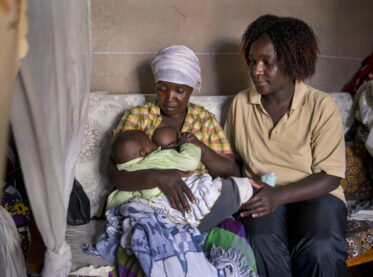 A community health worker sits on a sofa with a young mother as she breastfeeds her baby.